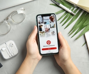 Someone holds a cellphone and looks at Pinterest. On the table is a palm frond, clear glasses and earpods.